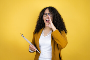 African american business woman with paperwork in hands over yellow background shouting and...