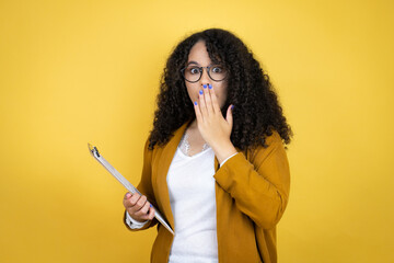 African american business woman with paperwork in hands over yellow background covering mouth with...