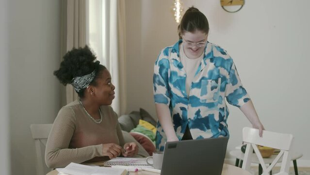 Medium shot of two ethnically diverse female roommates studying together for university in modern apartment, sitting at table with laptop, study materials and handmade sandwichesMedium shot of two eth