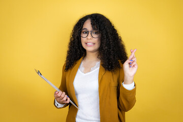 African american business woman with paperwork in hands over yellow background gesturing finger crossed smiling with hope and looking side - obrazy, fototapety, plakaty