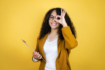 African american business woman with paperwork in hands over yellow background doing ok gesture shocked with smiling face, eye looking through fingers