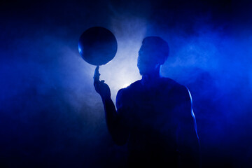 Basketball player silhouette lit with blue color spinning a ball against smoke background. Muscular african american man.