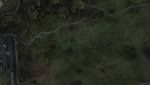 Aerial top view of park during dim twilight, featuring pathways, parking lot, grassy lawn and various trees. Space for text. Zoom out