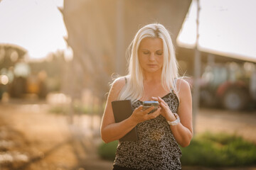 Valmiera, Latvia - August 17, 2024 - A focused blonde woman is holding a smartphone and a book on a farm with farming equipment in the background.