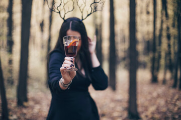 Mysterious woman in autumn forest wearing gothic deer antlers headband holding out mulled wine