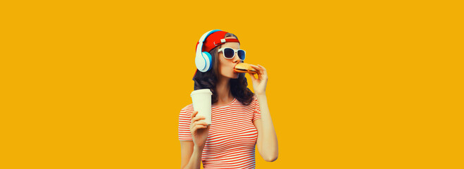 Stylish young woman listening to music in headphones eating burger fast food on yellow background