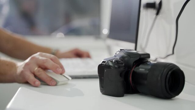 Professional black camera on desk, photographer's hands working with computer, uploading photo at the office. Modern photography equipment. Transferring photos from camera to computer. Close-up.