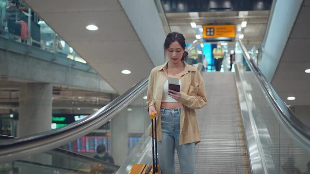 Asian tourist woman traveller with with a yellow suitcase using mobile phone while stand on moving walkway in airport terminal, Tourist journey trip concept.