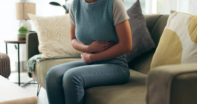 Hands, stomach pain and woman on sofa in living room with menstruation, gas or constipation at home. Gut health, ibs and female person with tummy ache for pms, nausea or endometriosis at apartment.