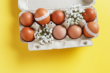 Brown eggs decorated with lace and white gypsophila flowers in carton on yellow background. Easter...