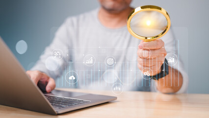 A man is holding a magnifying glass over a laptop computer. The laptop screen shows a variety of icons and symbols, including a clock, a smiley face, and a dollar sign, graph