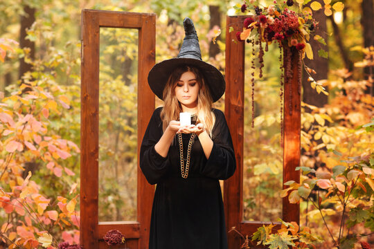 Young woman in Witch costum and hat with candle in hands stands against autumn forest background