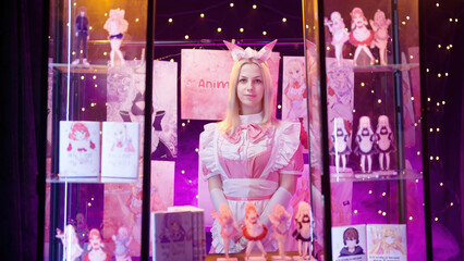 Cute pink cosplay maid girl selling anime art and figures on a stand