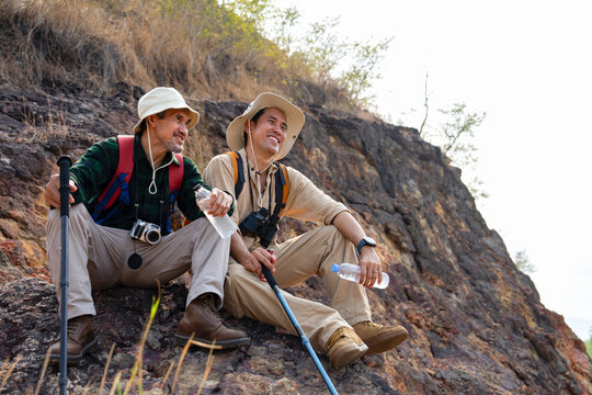 asian senior man with young adult male resting while climbing up steep on hill,happy father and adult son hiking together on summer time,concept of family lifestyle,active,adventure,journey