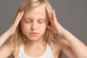 Fototapeta premium headache. little girl with long blonde hair stands on a gray background and holds her head with her hands, close-up, illness concept