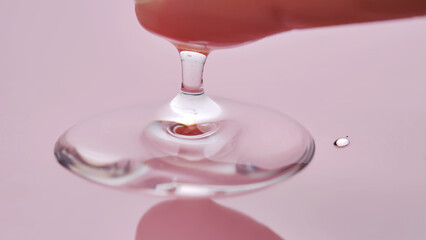 A finger touches a drop of cosmetic product on a pink background.