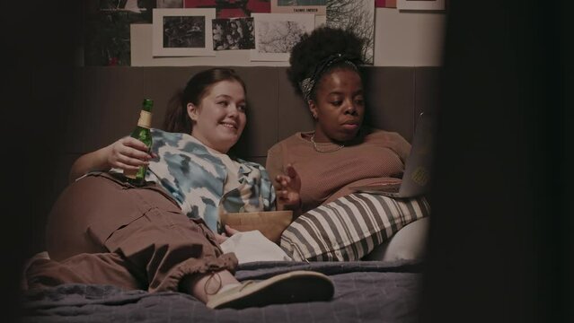 Two young multiethnic female best friends and roommates watching movie on laptop and chatting while lying in cozy bed in their apartment at night