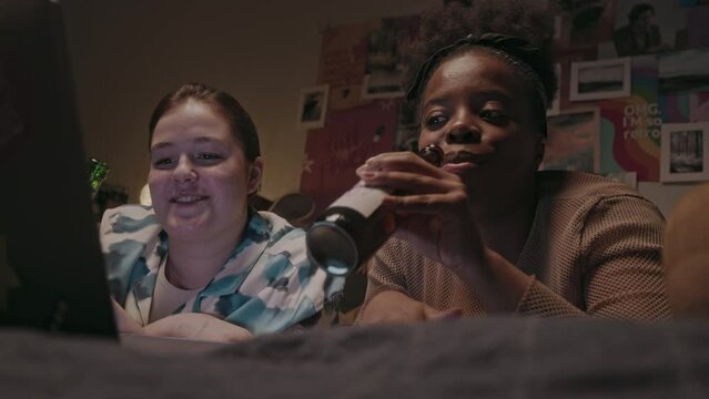 Chest up of two young multiracial girlfriends drinking cider and chatting while watching movie on laptop computer during pajama party sleepover