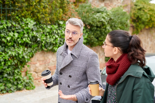 Business people in trendy outlooks and glasses walking down the street with takeaway coffee in hands and discussing the latest news or agenda in an informal setting.