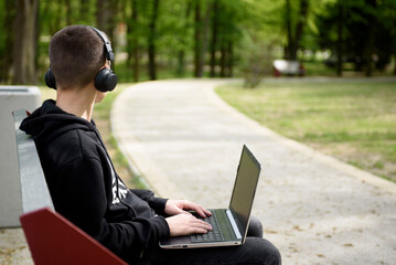  A young guy in headphones sits on a bench in the park with a laptop. Zielona Gora, Poland - 04.13.2024, Millennium Park.  