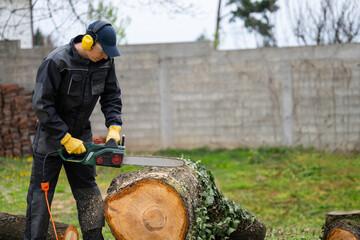A man in uniform cuts an old tree in the yard with an electric saw