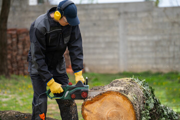 A man in uniform cuts an old tree in the yard with an electric saw - obrazy, fototapety, plakaty
