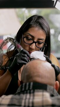 Panning of a female tattoo artist doing a scalp on a man's head, inside her studio