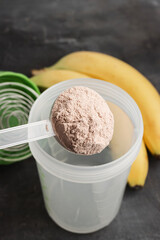 Plastic measuring spoon puts whey protein powder into a shaker on a dark background with bananas, process of making healthy protein drink