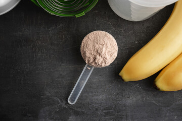 Whey protein powder with a chocolate flavor in a plastic measuring spoon on a dark background with bananas and a shaker, top view