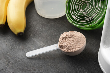 Whey protein powder with a chocolate flavor in a plastic measuring spoon on a dark background with bananas and a shaker