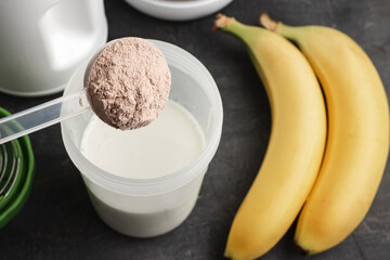 Plastic measuring spoon puts whey protein powder into a shaker with milk on a dark background with bananas, process of making healthy protein drink