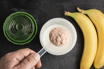 Male hand with a plastic measuring spoon puts whey protein powder into a shaker with milk on a dark background with bananas, process of making healthy protein drink, top view