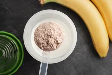 Plastic measuring spoon puts whey protein powder into a shaker with milk on a dark background with bananas, process of making healthy protein drink, top view