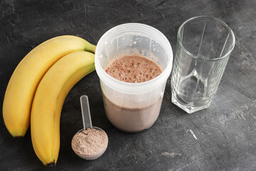 Blended chocolate protein drink in a shaker, plastic measuring spoon with protein powder, bananas...