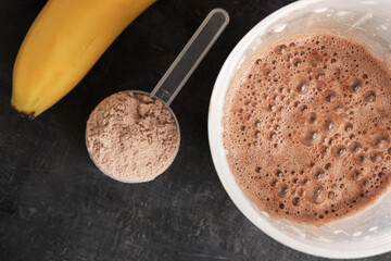 Blended chocolate protein drink in a shaker, plastic measuring spoon with protein powder, bananas on a dark background, top view