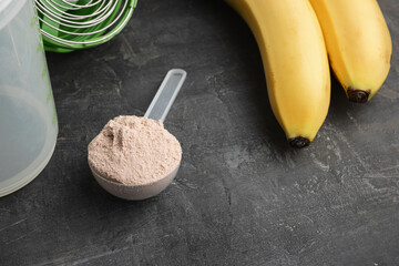 Whey protein powder with a chocolate flavor in a plastic measuring spoon on a dark background with bananas and a shaker