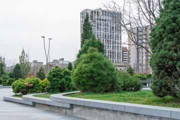 Nice modern leisure city park with benchs surrounded high-rise building. Public park in town residential. Sidewalk and walkway with conifer trees. Place to rest in city landscape. Urban architecture.