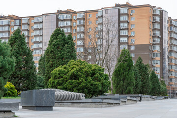 Nice modern leisure city park with benchs surrounded high-rise building. Public park in town residential. Sidewalk and walkway with conifer trees. Place to rest in city landscape. Urban architecture.