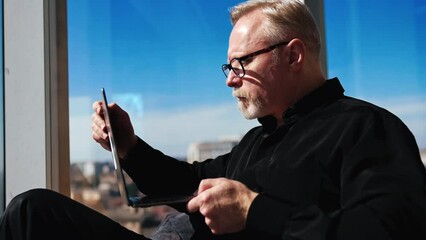 Concentrated male in black outfit sits ay window holding a laptop. Man in glasses checks e-mail and closes his gadget.