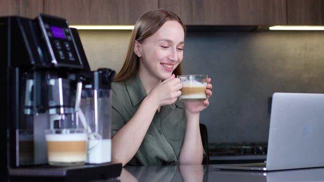 Woman drinking coffee and working on laptop near coffee machine at home
