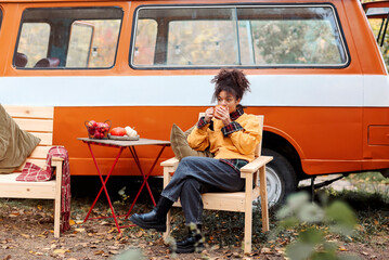 Young african american woman sitting at table near car in autumn forest and drinking hot tea - Powered by Adobe