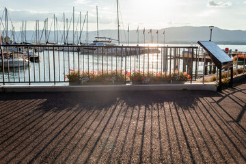 Sunlight filters through a marina on Lake Leman, casting long shadows on the pavement, while boats...