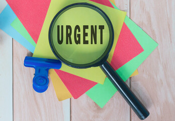Urgent word on colorful adhesive paper with magnifying glass on wooden table.