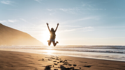 Happy woman with backpack jumping with arms raised on the beach at sunset - Lovely tourist enjoying summer vacation by the sea - Traveler lifestyle and wellness concept