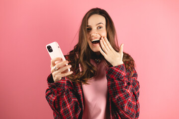 Shocked brunette woman wear plaid shirt hold mobile phone isolated on pink background studio....