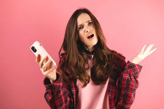 Mad and shock young brunette woman using mobile phone isolated on pink background. Yelling unhappy girl hold mobile phone, raised hands. Disappointed sad upset lady horrified impressed news. WTF Oh no