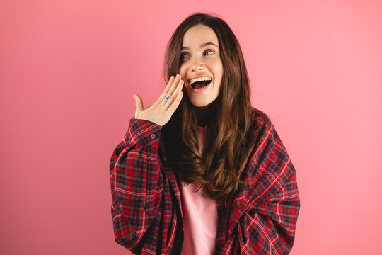 Wow! Cheerful young brunette woman, amazed, covering mouth with hand, standing over pink isolated background. Beautiful expression of happy surprise, look at side, gossip girl!