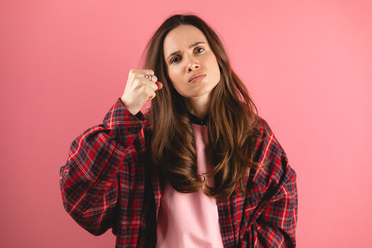 Brunette woman threatens with a fist is isolated on pink background, shows fist has annoyed face expression going to revenge or threaten someone makes serious look. I will show you who is boss.