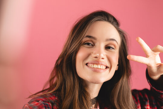 Close up photo of beautiful young brunette woman taking selfie, posing with peace v-sign, smiling happy, take photo, straight hand to camera posing against pink background.