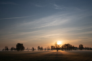 Landscape in Bavaria. The sun rises behind bare trees. There are soft clouds in the sky. Against...